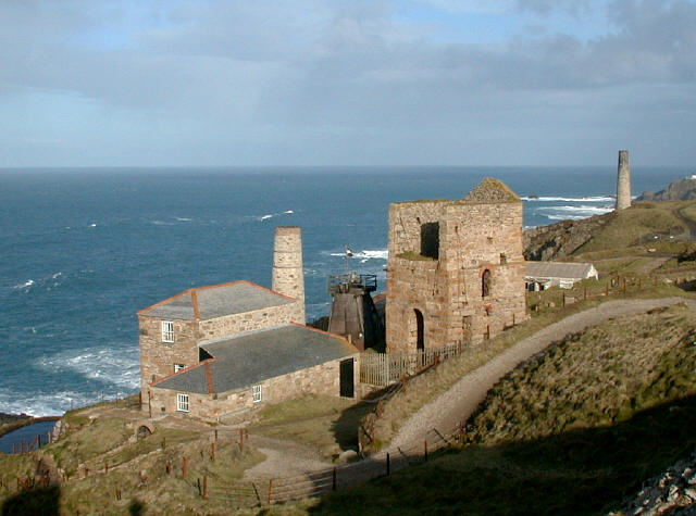 A ruin of a pump house with two other restored buildings, one with chimney. The group is at the top of a sea cliff
